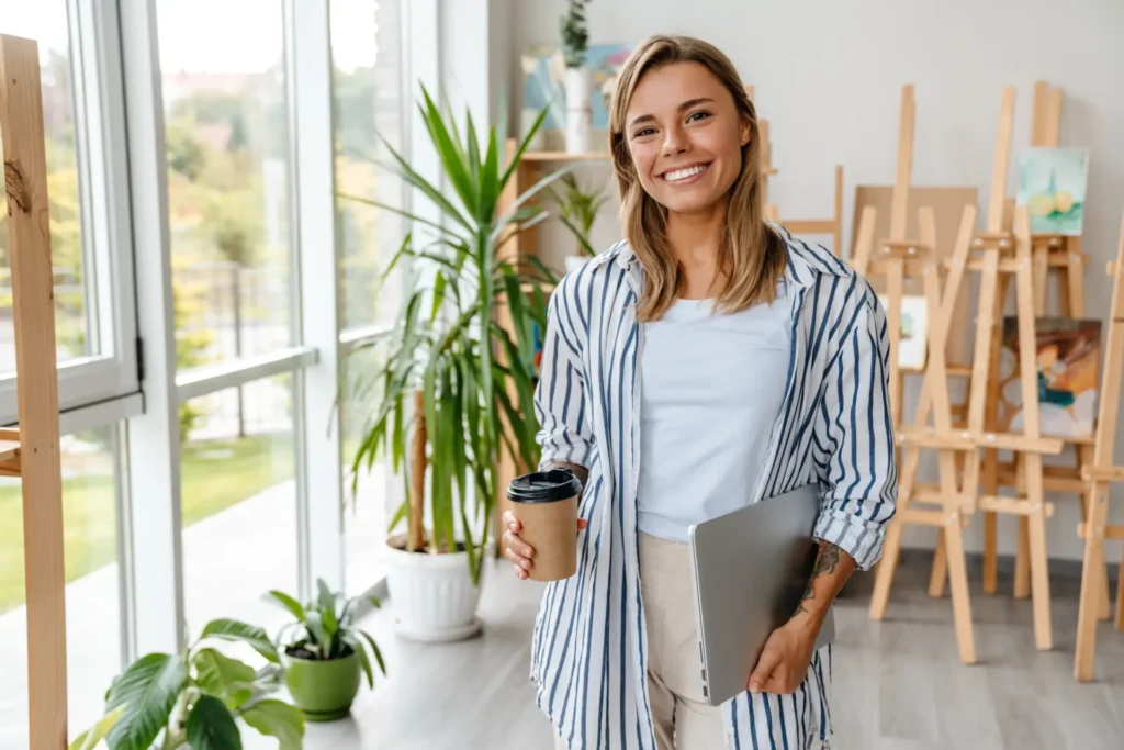 Lächelnde Frau mit Laptop und Kaffeebecher in einem Innenraum mit Pflanzen und Staffeleien.
